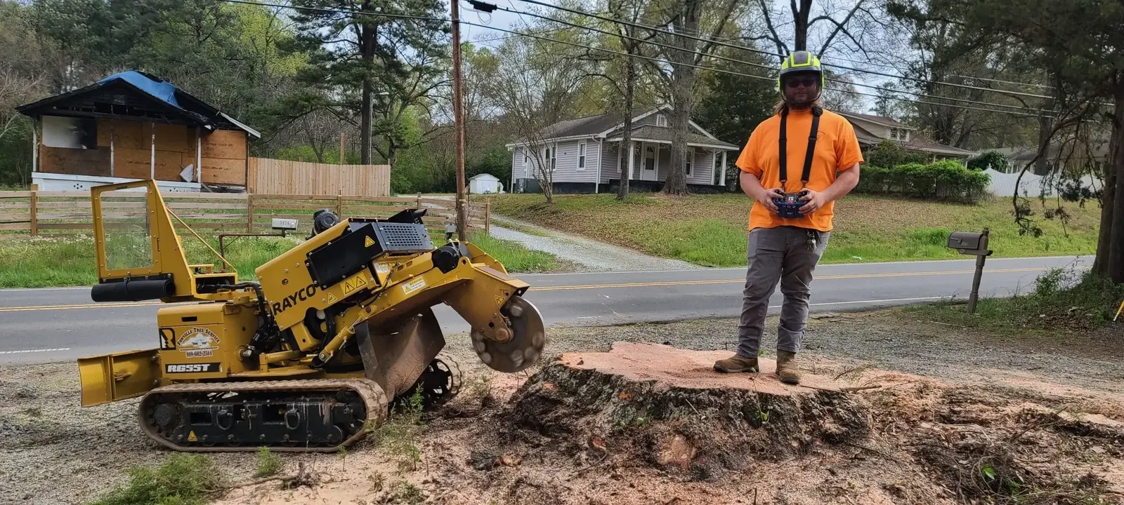 Fonvilletreeservice crew member operating remote-controlled Rayco stump grinder on large tree stump in residential NC neighborhood.